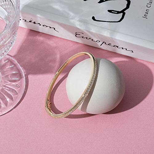 Elegant gold bangle resting on a round white object with a book and glass nearby on a pink surface.